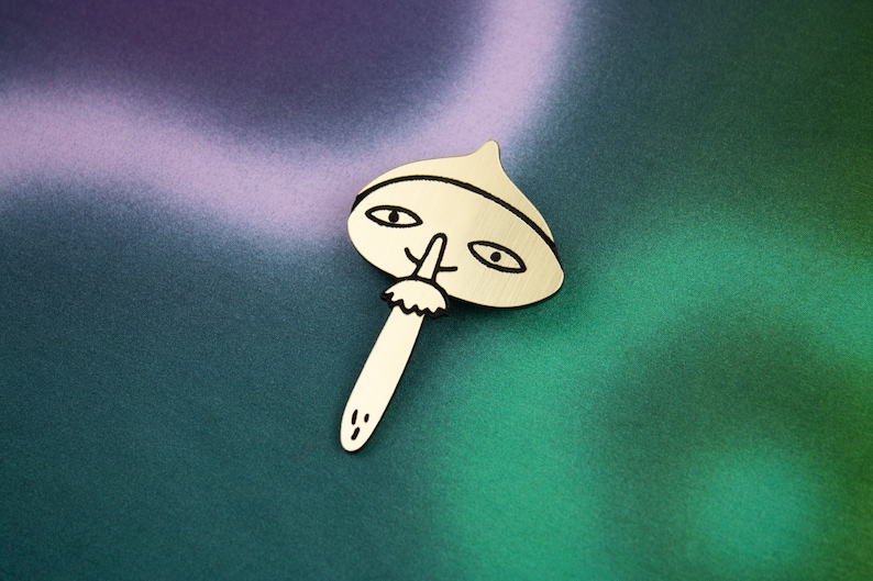 May include: A gold-coloured metal pin shaped like a mushroom with a face. The mushroom has a black outline and black eyes, a nose, and a moustache.