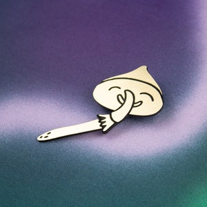 May include: A gold-coloured metal pin shaped like a mushroom with a smiling face and a hand covering its mouth. The mushroom is lying on a purple and green background.