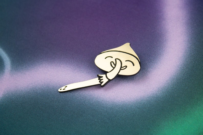 May include: A gold-coloured metal pin shaped like a mushroom with a smiling face and a hand covering its mouth. The mushroom is lying on a purple and green background.
