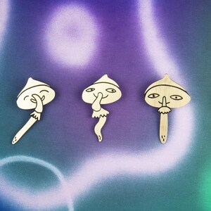May include: Five wooden mushroom figurines with different facial expressions. Each mushroom has a white painted body and black painted details. The mushrooms are arranged in a row on a purple and green background.