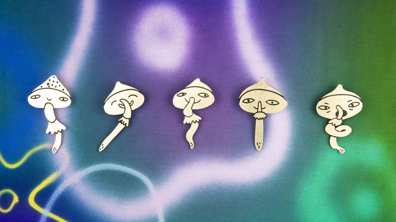 May include: Five wooden mushroom figurines with different facial expressions. Each mushroom has a white painted body and black painted details. The mushrooms are arranged in a row on a purple and green background.