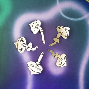 May include: Five silver pins shaped like mushrooms with whimsical faces and worm-like appendages. The pins are arranged in a circle on a purple and green tie-dye background.