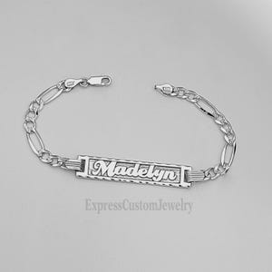 May include: Silver bracelet with a personalized nameplate that reads "Madelyn". The bracelet features a delicate figaro chain.