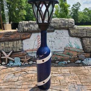 May include: A decorative solar-powered garden lamp made from a repurposed blue wine bottle. The bottle is painted with a white stripe and lighthouse design, with a black lamp fixture on top. The bottle is decorated with white specks and small bird silhouettes.