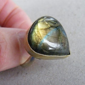 May include: A handcrafted ring featuring a large, triangular labradorite gemstone. The iridescent stone displays shades of blue, green, and gold. The gemstone is set in a gold-colored metal band, creating a unique and eye-catching piece of jewelry.