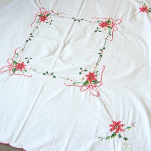 Könnte beinhalten: Weißes Tischtuch mit gestickten roten Weihnachtssternen, grünen Stechpalmenblättern und rosa Bändern. Die Ränder haben eine rosa gewellte Bordüre. Ein festliches Textil.