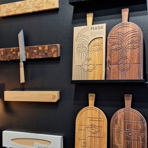 May include: A display of wooden cutting boards with various designs. Some boards feature a carved face design, while others have a more traditional look. The boards are displayed on a black wall with a magnetic knife holder and a knife.