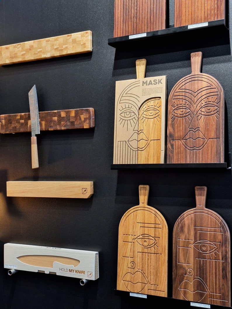 May include: A display of wooden cutting boards with various designs. Some boards feature a carved face design, while others have a more traditional look. The boards are displayed on a black wall with a magnetic knife holder and a knife.