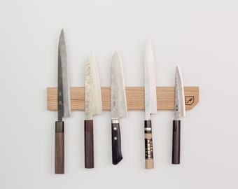 Peut inclure: Cinq couteaux de cuisine avec des manches en bois sont magnétiquement fixés à une bande magnétique en bois montée sur un mur blanc. Les couteaux sont disposés en rangée de gauche à droite, le plus long couteau à gauche et le plus court couteau à droite.