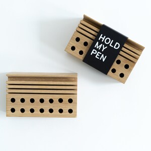 May include: Two wooden pen holders with a black label on one that says "HOLD MY PEN". Both holders have multiple holes for pens and pencils.