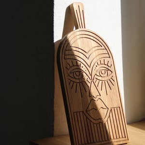 May include: A wooden cutting board with a carved face design. The face has large eyes, a nose, and a mouth. The board is made of light brown wood and has a natural grain.