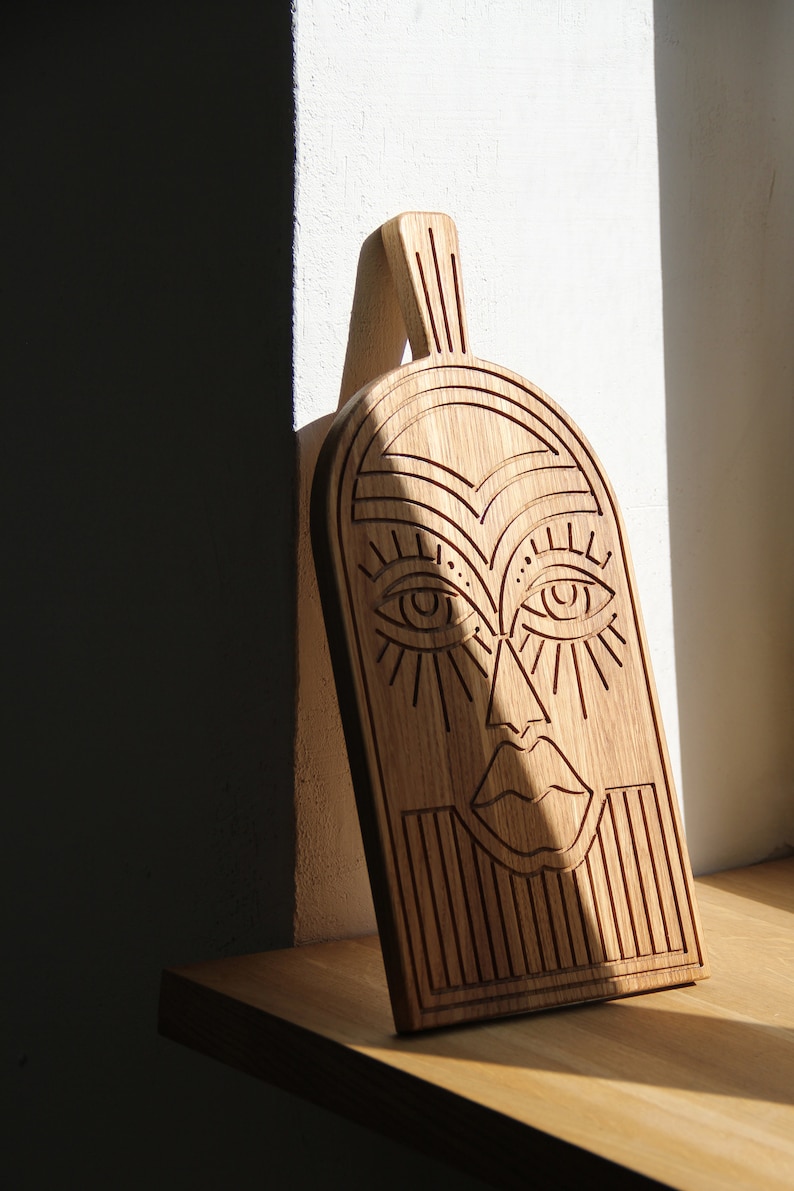 May include: A wooden cutting board with a carved face design. The face has large eyes, a nose, and a mouth. The board is made of light brown wood and has a natural grain.