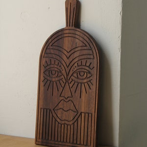 May include: A wooden cutting board with a carved design of a face. The face has large eyes, a nose, and a mouth. The board is dark brown and has a smooth finish.