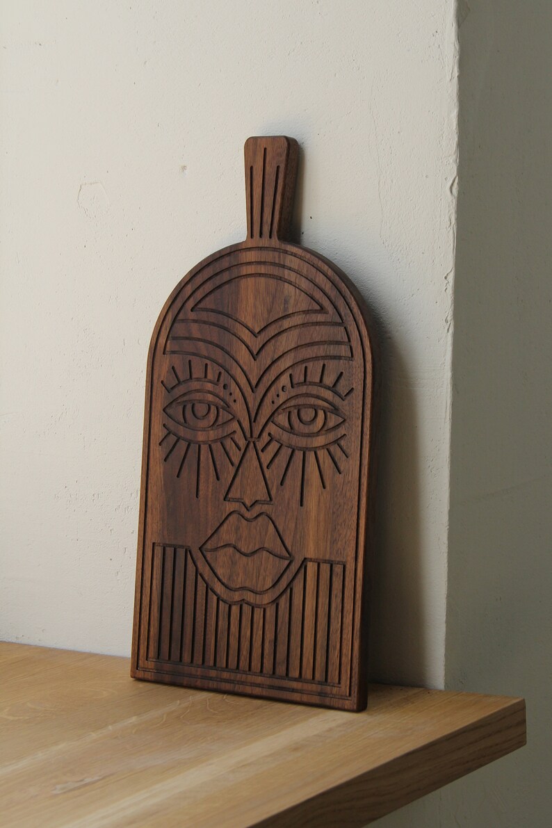 May include: A wooden cutting board with a carved design of a face. The face has large eyes, a nose, and a mouth. The board is dark brown and has a smooth finish.