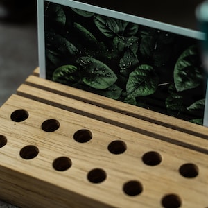 May include: A wooden organizer with multiple slots for pens and pencils. The organizer has a slot for a photo or card, which is displaying a photo of green leaves. A blue pen is also in the organizer.