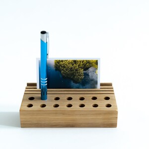 May include: A wooden desk organizer with multiple slots for pens and pencils. A blue pen is in one of the slots and a photo is in the top slot.