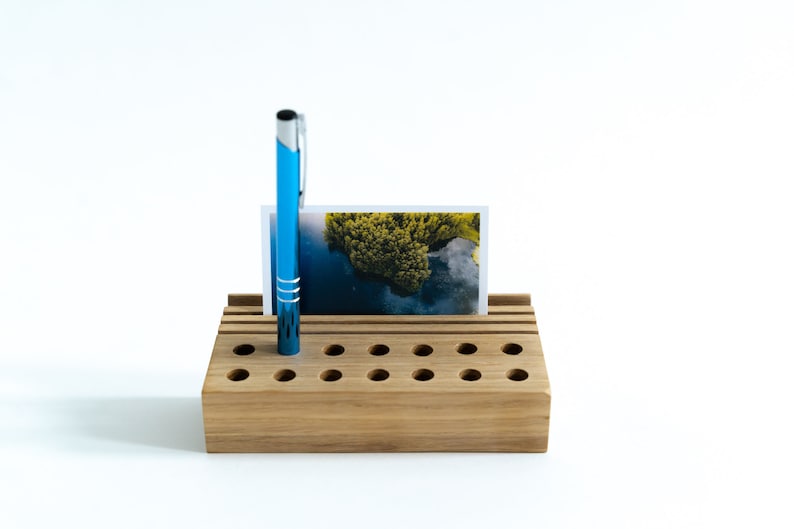 May include: A wooden desk organizer with multiple slots for pens and pencils. A blue pen is in one of the slots and a photo is in the top slot.