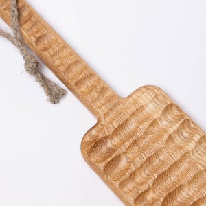 May include: A long, wooden serving tray with a rope handle. The tray has a textured surface and is made of light brown wood.