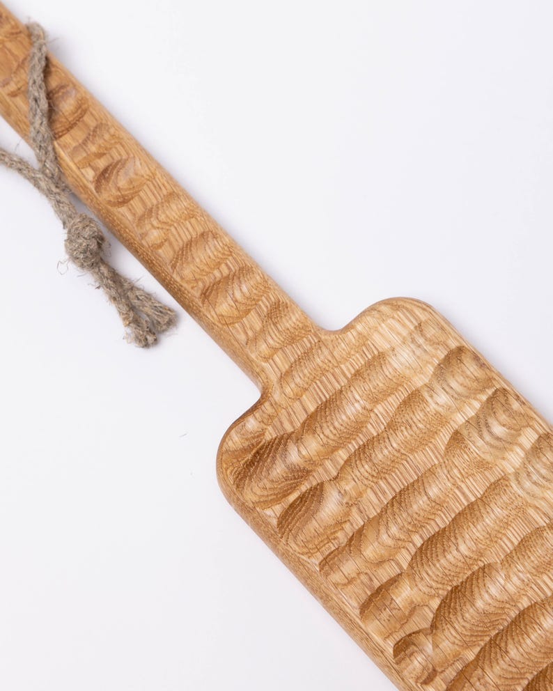 May include: A long, wooden serving tray with a rope handle. The tray has a textured surface and is made of light brown wood.