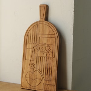 May include: A wooden cutting board with a carved abstract face design. The face features a large eye, a nose, and a mouth.
