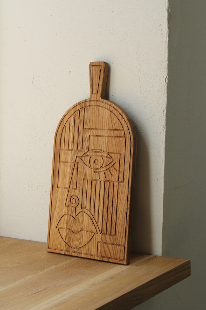 May include: A wooden cutting board with a carved abstract face design. The face features a large eye, a nose, and a mouth.
