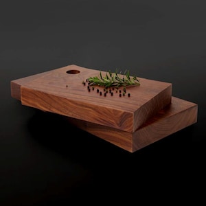 May include: Two wooden cutting boards, one stacked on top of the other. The top board has a small hole in the center and is decorated with a sprig of rosemary and black peppercorns.