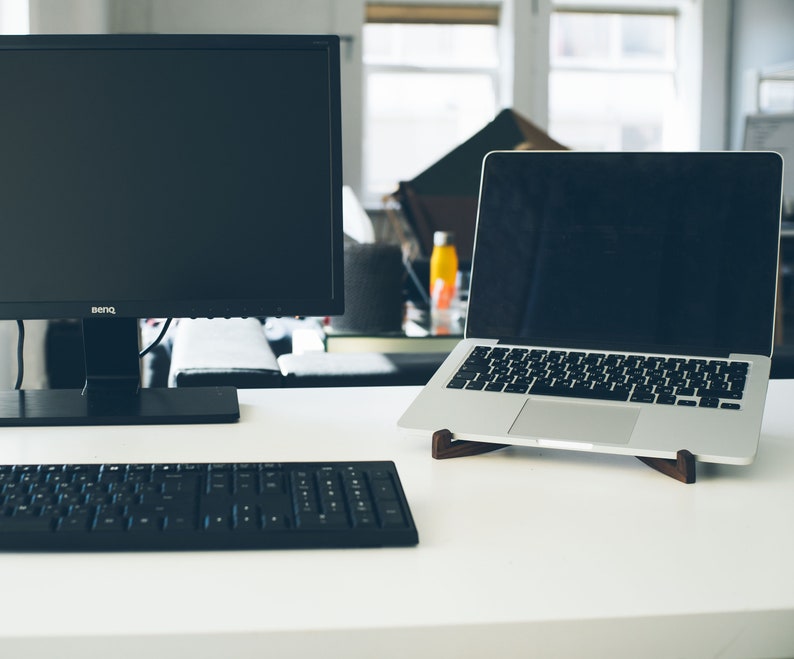 May include: A black computer monitor with a black keyboard and a silver laptop on a white desk. The laptop is on a wooden stand.