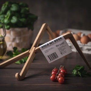 以下が含まれることがあります： 白いラベルに「HOLD MY BOOK WOODEN LEG FOR RECIPE BOOKS」というテキストとブランド名「Dir Lahr」が書かれた木製の本立て。スタンドは、赤いトマトの房、緑のパセリの枝、ニンニクの球根が置かれた暗い木製の表面の上に置かれています。