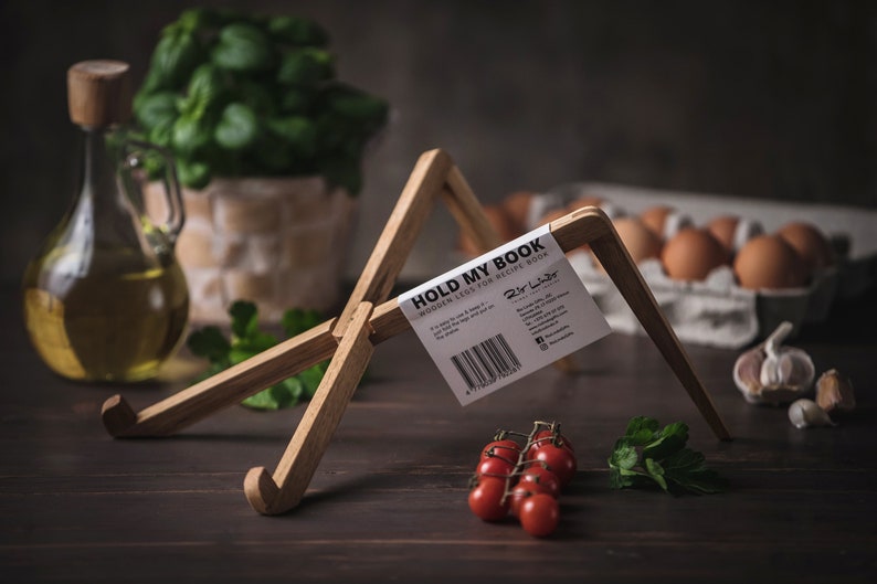 以下が含まれることがあります： 白いラベルに「HOLD MY BOOK WOODEN LEG FOR RECIPE BOOKS」というテキストとブランド名「Dir Lahr」が書かれた木製の本立て。スタンドは、赤いトマトの房、緑のパセリの枝、ニンニクの球根が置かれた暗い木製の表面の上に置かれています。