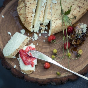 Puede incluir: Una tabla de cortar de madera redonda con forma de &aacute;rbol, con un queso blanco con corteza marr&oacute;n, cortado en rodajas y dispuesto en la tabla. Un peque&ntilde;o cuchillo se coloca en la tabla, y un peque&ntilde;o plato de miel es visible en el fondo. La tabla est&aacute; decorada con peque&ntilde;as bayas rojas y hojas verdes.