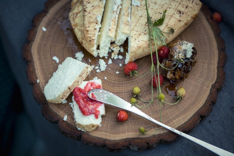 Puede incluir: Una tabla de cortar de madera redonda con forma de &aacute;rbol, con un queso blanco con corteza marr&oacute;n, cortado en rodajas y dispuesto en la tabla. Un peque&ntilde;o cuchillo se coloca en la tabla, y un peque&ntilde;o plato de miel es visible en el fondo. La tabla est&aacute; decorada con peque&ntilde;as bayas rojas y hojas verdes.