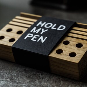 May include: A wooden pen holder with a black label that says "HOLD MY PEN". The holder has multiple slots for pens and pencils.