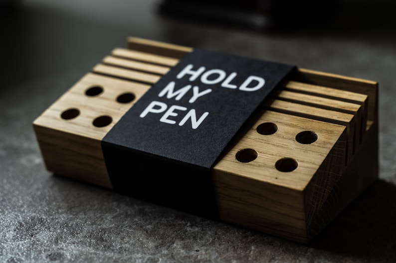 May include: A wooden pen holder with a black label that says "HOLD MY PEN". The holder has multiple slots for pens and pencils.
