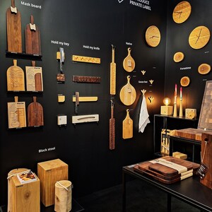 May include: A black wall display with various wooden products, including cutting boards, wall hooks, stools, and clocks. The text "Hand Made in Lithuania" and "We Produce Private Label" are visible on the wall.