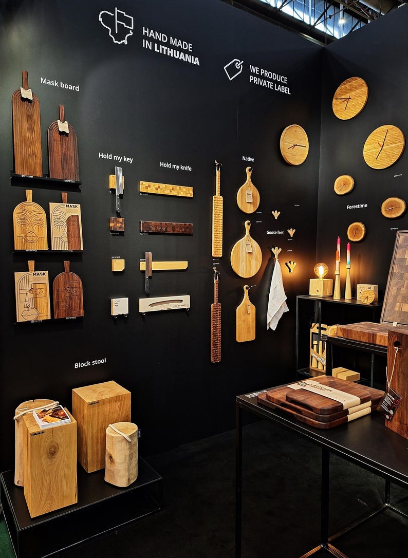 May include: A black wall display with various wooden products, including cutting boards, wall hooks, stools, and clocks. The text "Hand Made in Lithuania" and "We Produce Private Label" are visible on the wall.
