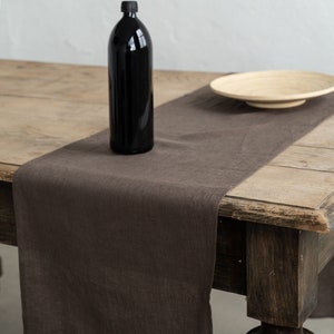 Könnte beinhalten: Ein brauner Tischläufer aus Leinen auf einem Holztisch mit einer schwarzen Glasflasche und einem Holzteller.