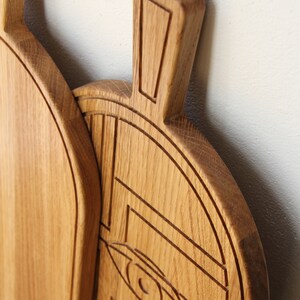 May include: A wooden cutting board with a carved design of an eye and geometric shapes. The board is made of light brown wood and has a natural finish.