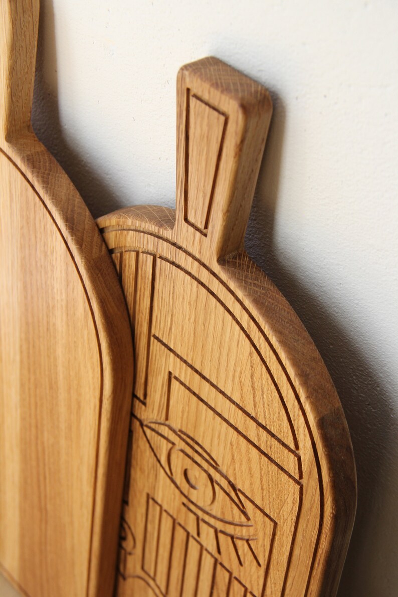 May include: A wooden cutting board with a carved design of an eye and geometric shapes. The board is made of light brown wood and has a natural finish.