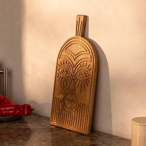 May include: A wooden cutting board with a carved face design. The board is rectangular with a handle and features detailed carvings of eyes, a nose, and lips. The wood has a natural, light brown color, and the board is leaning against a white wall.