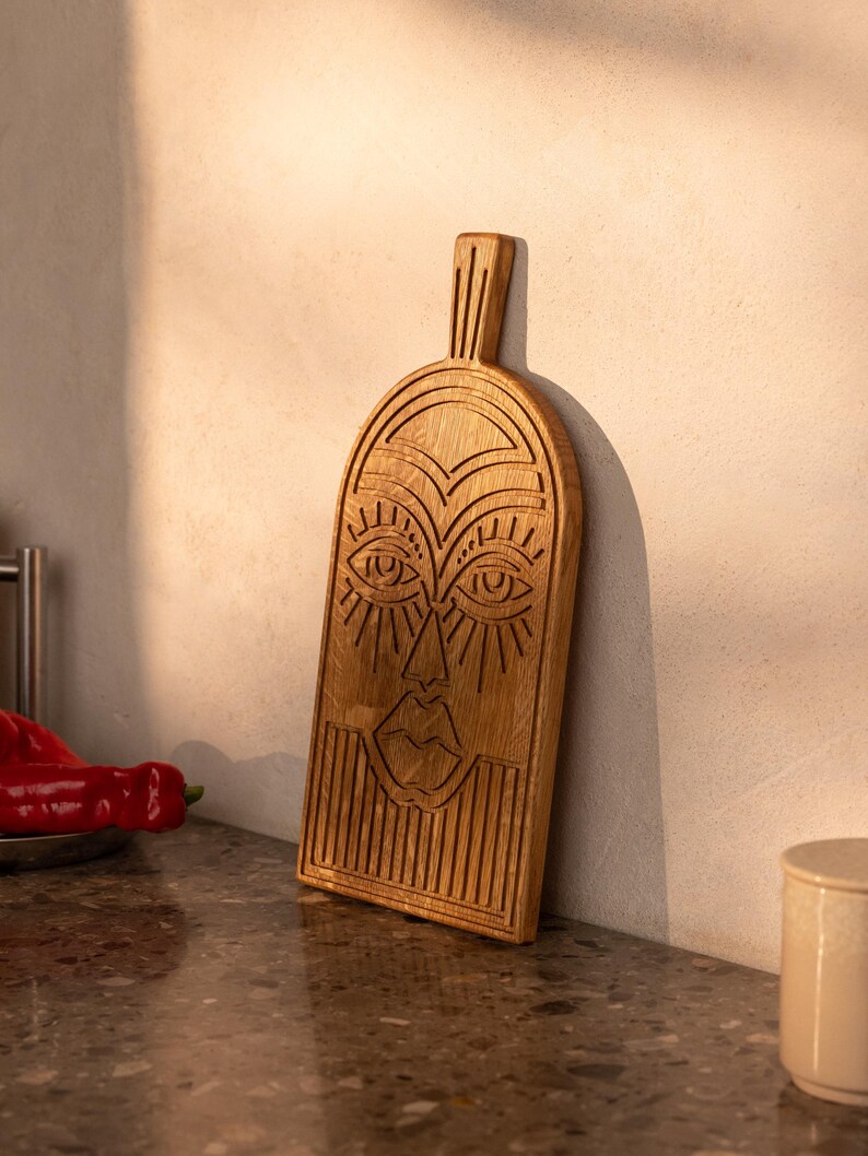 May include: A wooden cutting board with a carved face design. The board is rectangular with a handle and features detailed carvings of eyes, a nose, and lips. The wood has a natural, light brown color, and the board is leaning against a white wall.