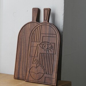 May include: Two wooden cutting boards with a geometric face design carved into the surface. The boards are dark brown and have a natural wood finish.