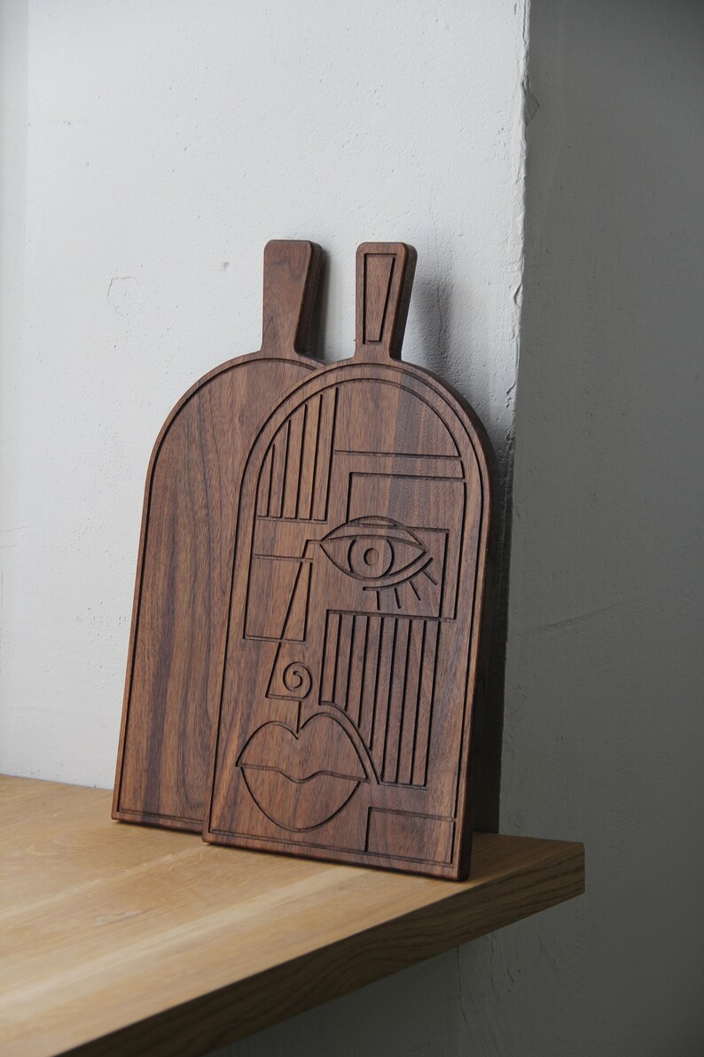 May include: Two wooden cutting boards with a geometric face design carved into the surface. The boards are dark brown and have a natural wood finish.