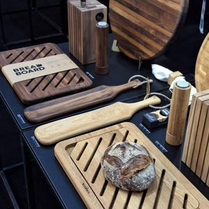 May include: A collection of wooden kitchenware including rectangular bread boards with diagonal slats, long serving paddles with rope loops, cylindrical salt and pepper mills, a knife block, and a round board. A loaf of bread sits on one board.