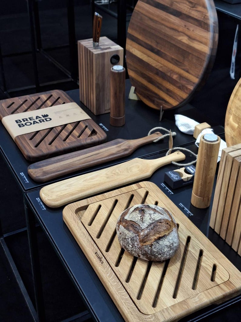 May include: A collection of wooden kitchenware including rectangular bread boards with diagonal slats, long serving paddles with rope loops, cylindrical salt and pepper mills, a knife block, and a round board. A loaf of bread sits on one board.