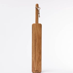 May include: A long, rectangular wooden cutting board with a rope handle. The board is made of light brown wood with a natural grain pattern. The board is labeled "woodlore" on the bottom.