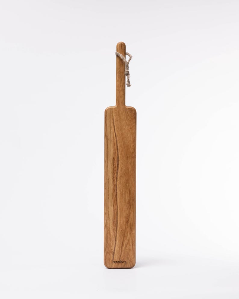 May include: A long, rectangular wooden cutting board with a rope handle. The board is made of light brown wood with a natural grain pattern. The board is labeled "woodlore" on the bottom.