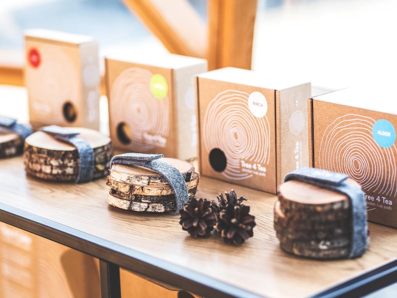 Puede incluir: Cuatro juegos de posavasos de madera con fondo de fieltro, cada juego apilado uno encima del otro. Los posavasos est&aacute;n hechos de diferentes tipos de madera, incluyendo abedul, aliso y roble. Cada juego est&aacute; empaquetado en una caja de cart&oacute;n con un dise&ntilde;o de veta de madera y el texto "Tree 4 Tea" impreso en la parte delantera.