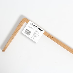 以下が含まれることがあります： 薄い茶色の木で作られた木製の本立て。白いラベルに「HOLD MY BOOK WOODEN LEAN FOR RECIPE BOOK」と「The Label」のロゴが印刷されています。
