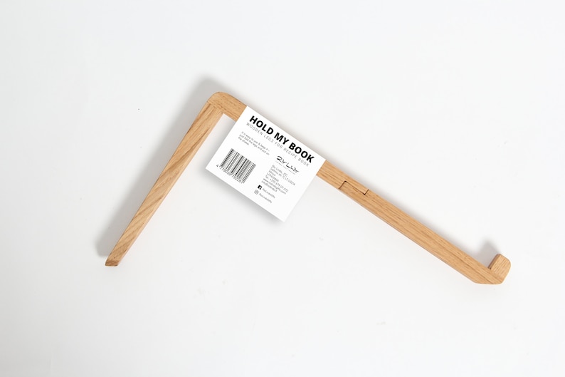 以下が含まれることがあります： 薄い茶色の木で作られた木製の本立て。白いラベルに「HOLD MY BOOK WOODEN LEAN FOR RECIPE BOOK」と「The Label」のロゴが印刷されています。