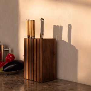 May include: A rectangular dark wood knife block featuring vertical slats and four slots. Three knives with light-colored wooden handles and one knife with a metallic handle are inserted into the block, which stands upright on a speckled surface.